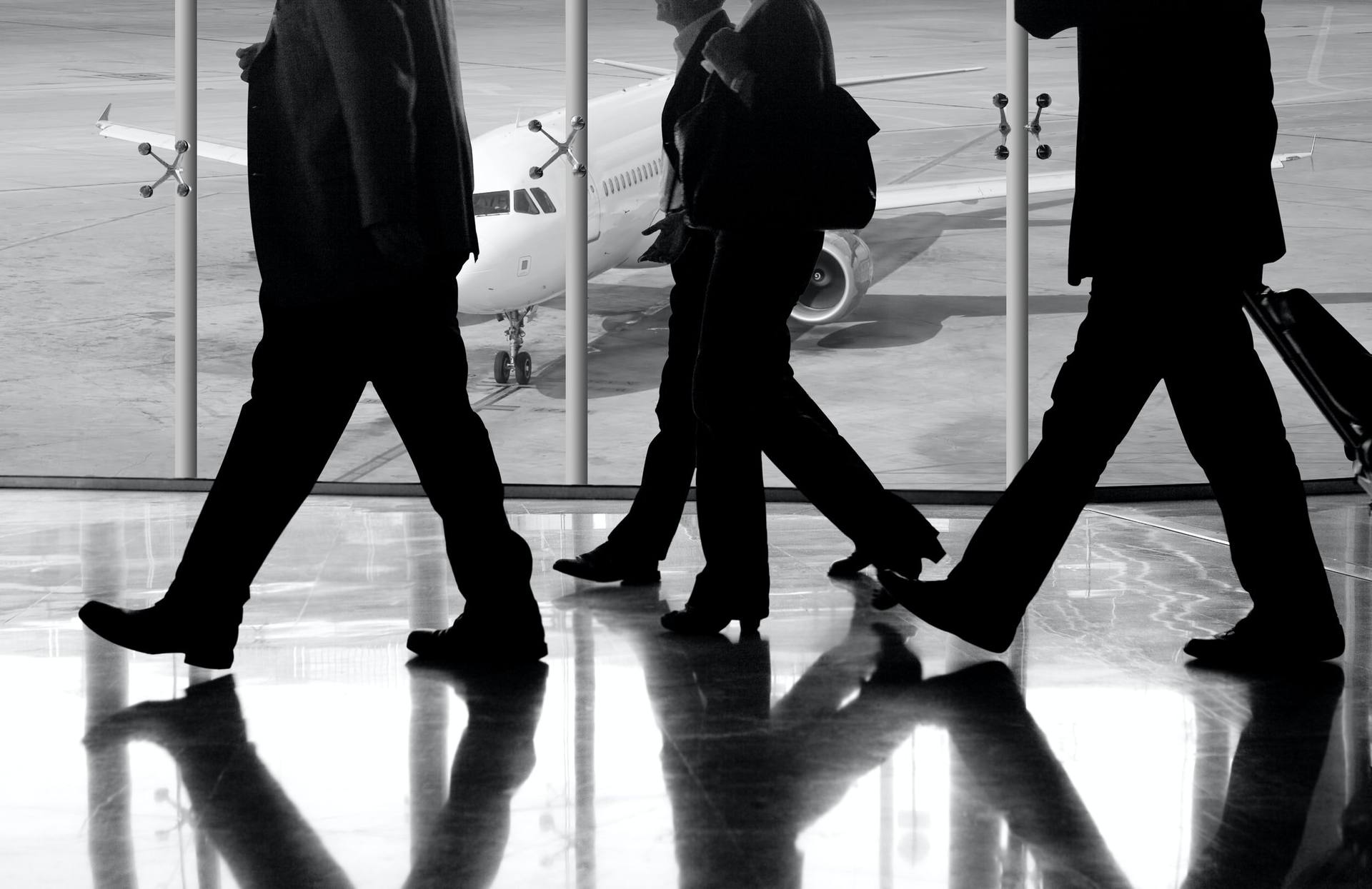 Commuters crossing a bright terminal hall in silhouette, an aircraft waiting on the tarmac beyond the glass